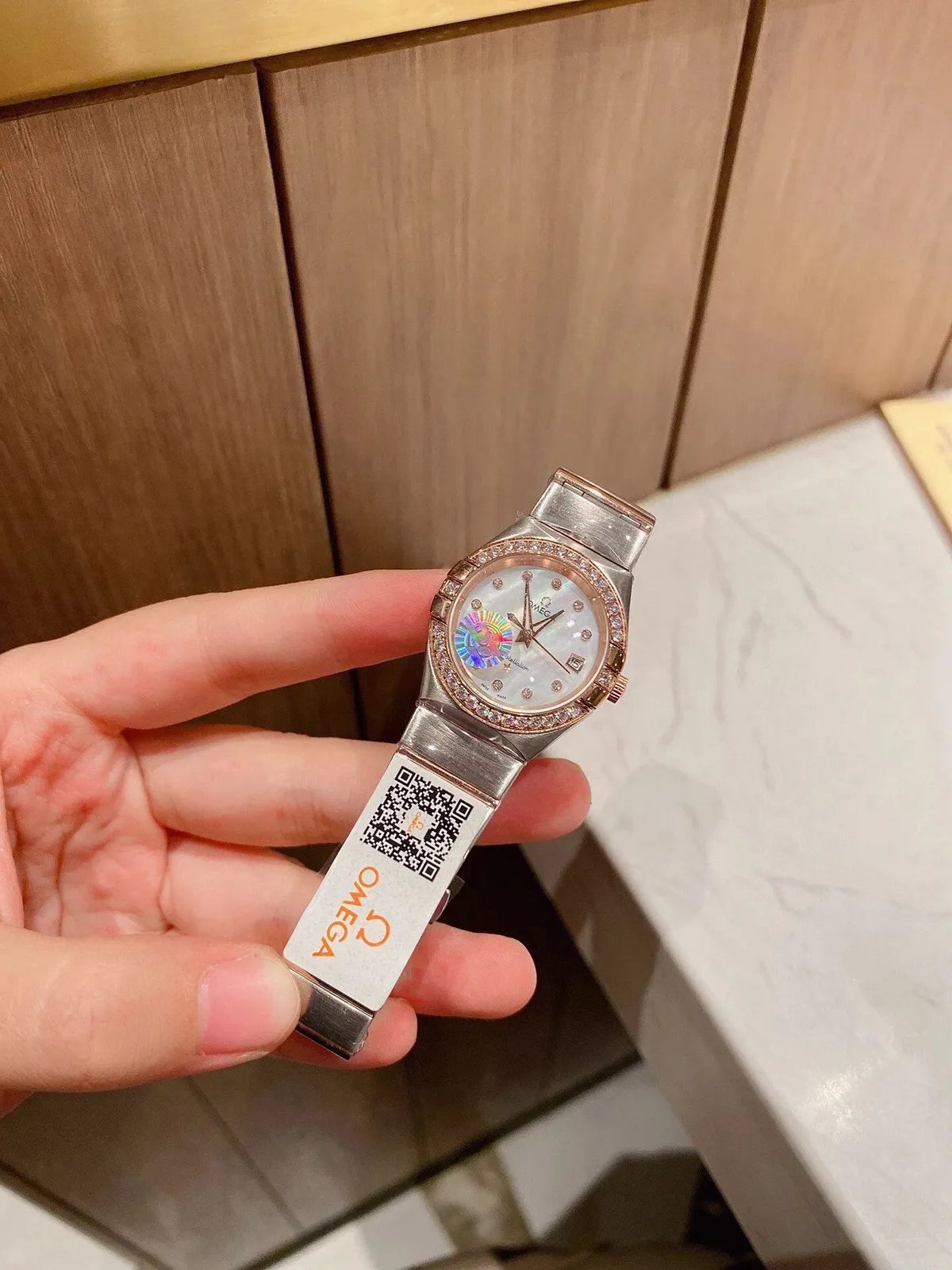 Omega zegarek damski z metalową bransoletą i kamienną ramką na jasnej tarczy.