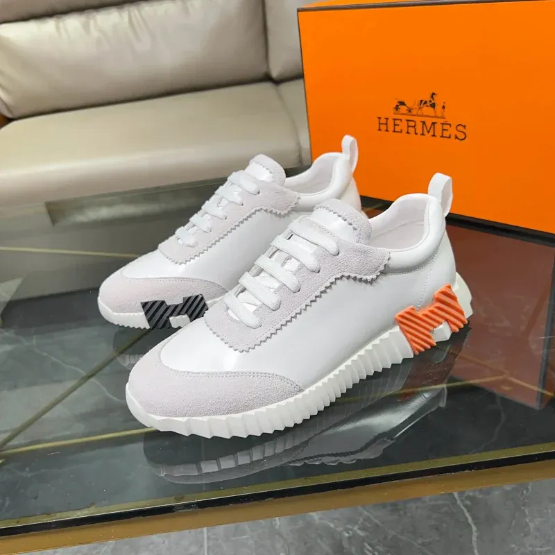 Hermes sneakersy niski profil w bieli, szarości i pomarańczu na szklanym stole.